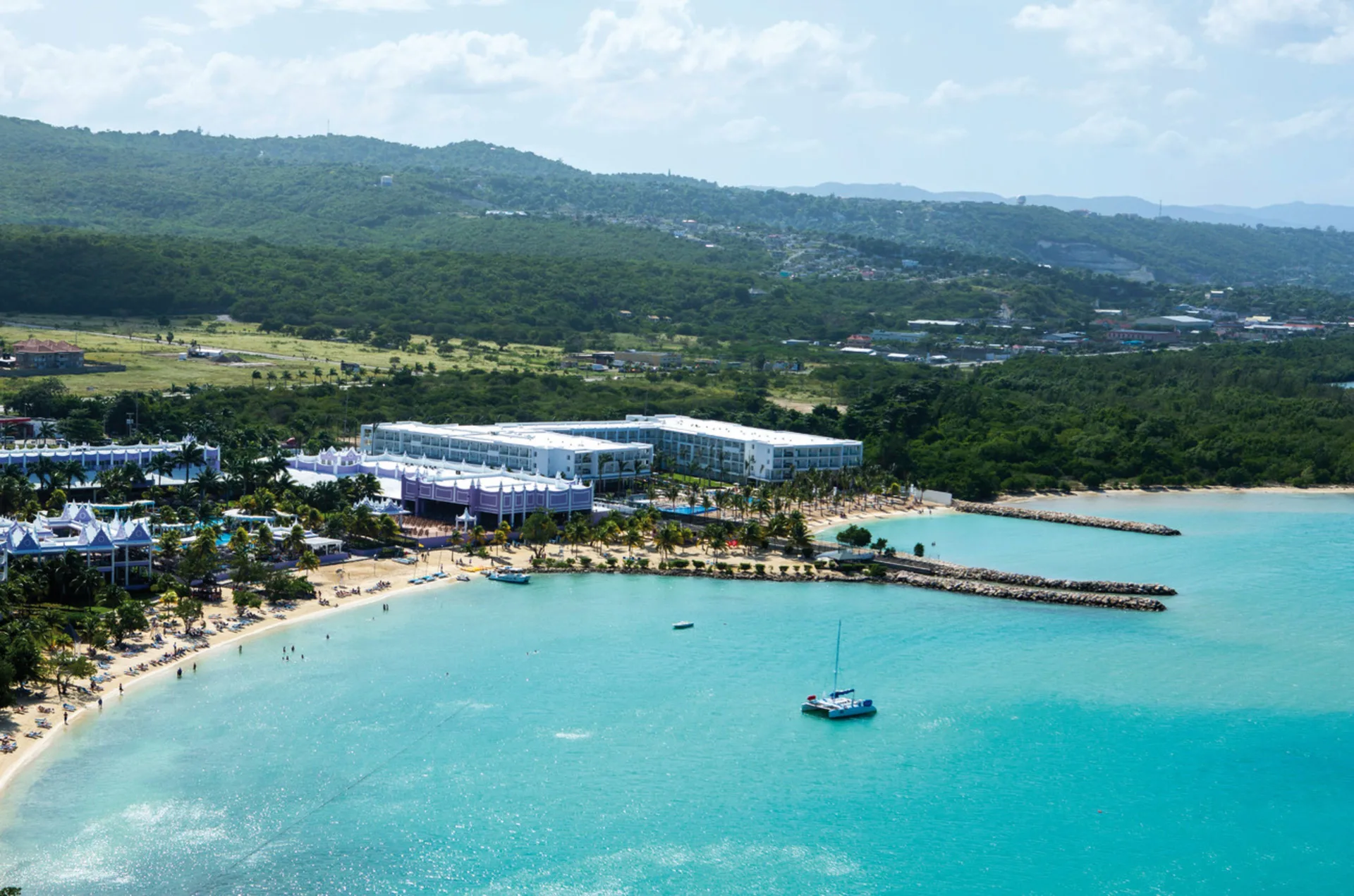 Wide aerial view of Hotel Riu Palace Jamaica on the bay with beach, hills, and a catamaran offshore.