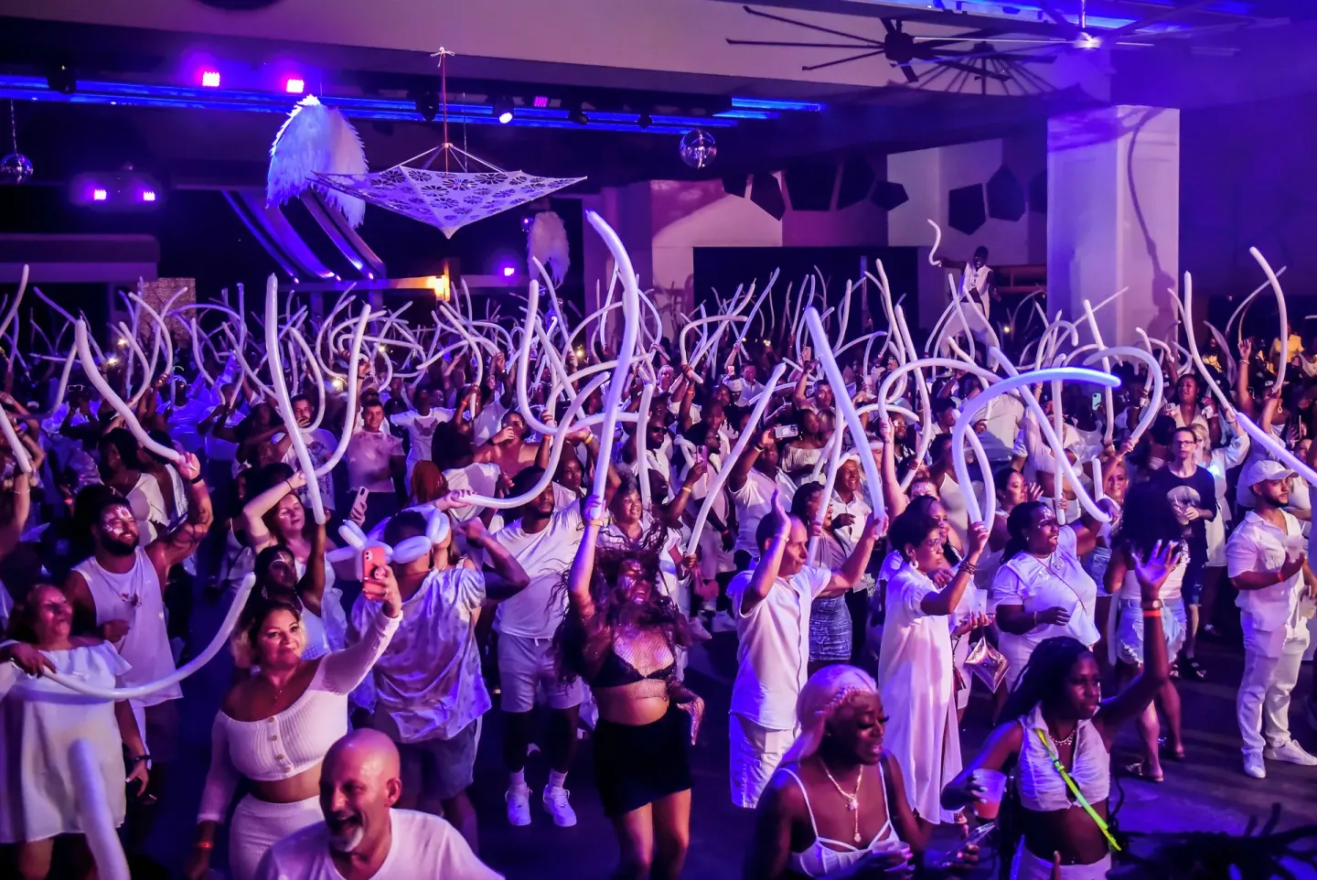 Crowd waves white balloons at a packed indoor party at Hotel Riu Palace Jamaica.