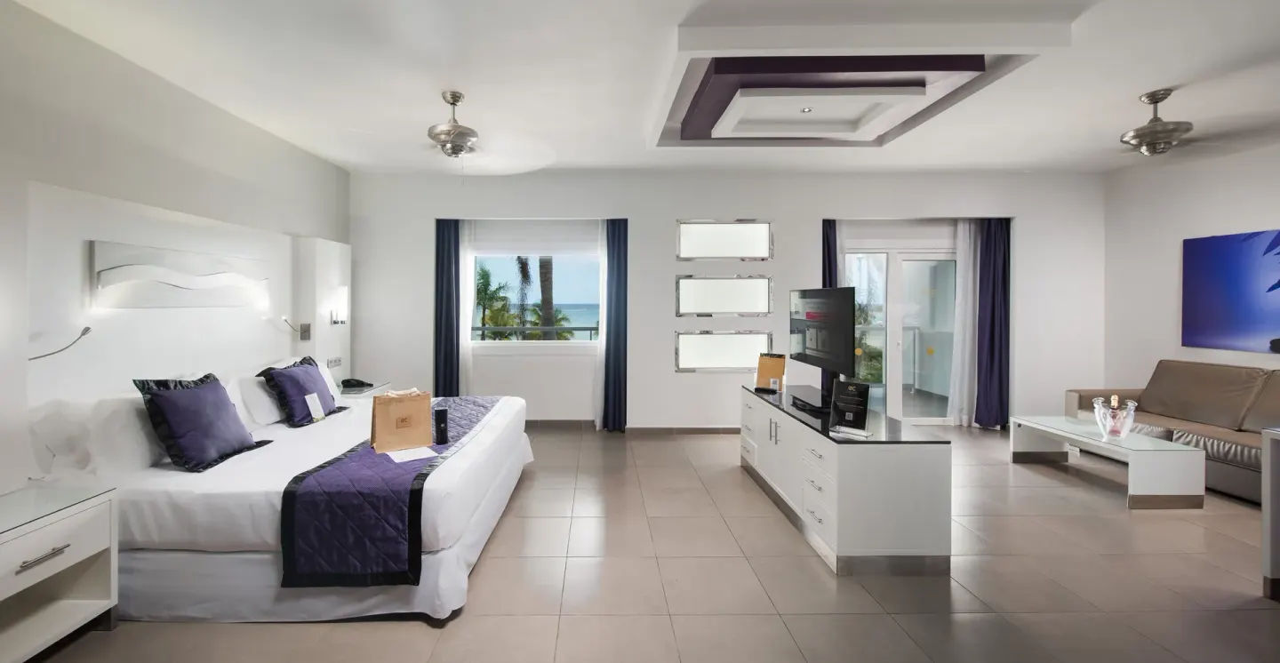 Guest suite with king bed, sofa, TV console, and ocean-view window at Hotel Riu Palace Jamaica.