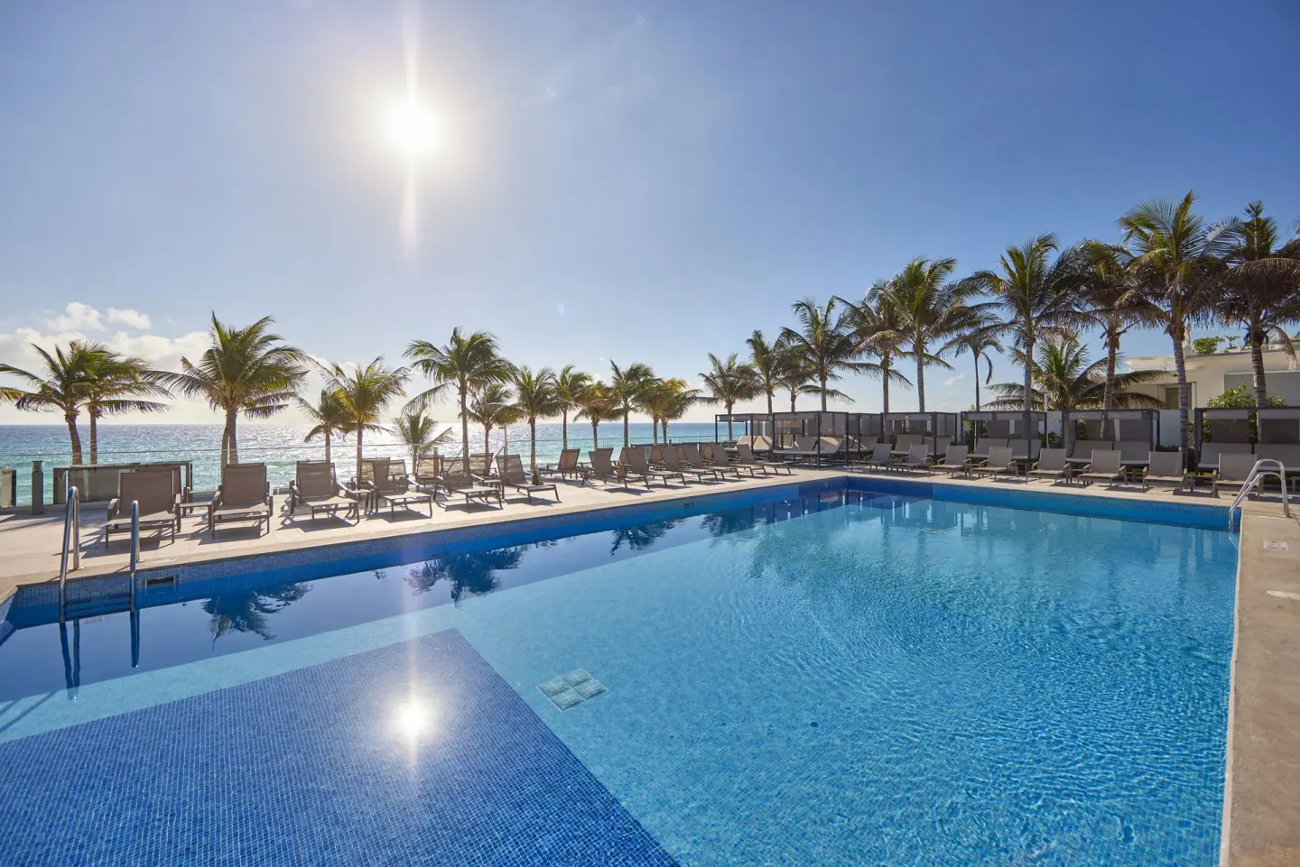 Oceanfront swimming pool with palm trees and loungers at Hotel Riu Palace Kukulkan.