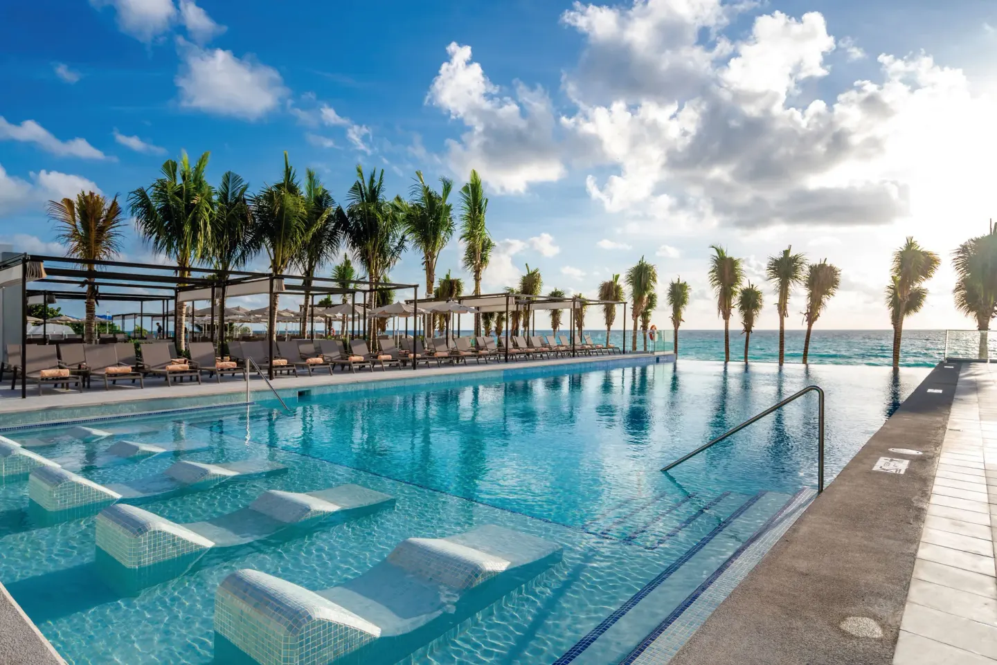 Pool deck with submerged loungers and ocean view at Hotel Riu Palace Kukulkan.