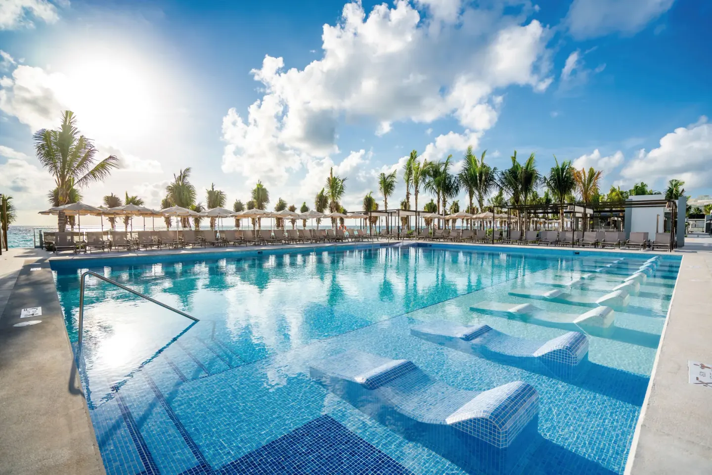 Sunny pool with submerged loungers and oceanfront deck at Hotel Riu Palace Kukulkan.