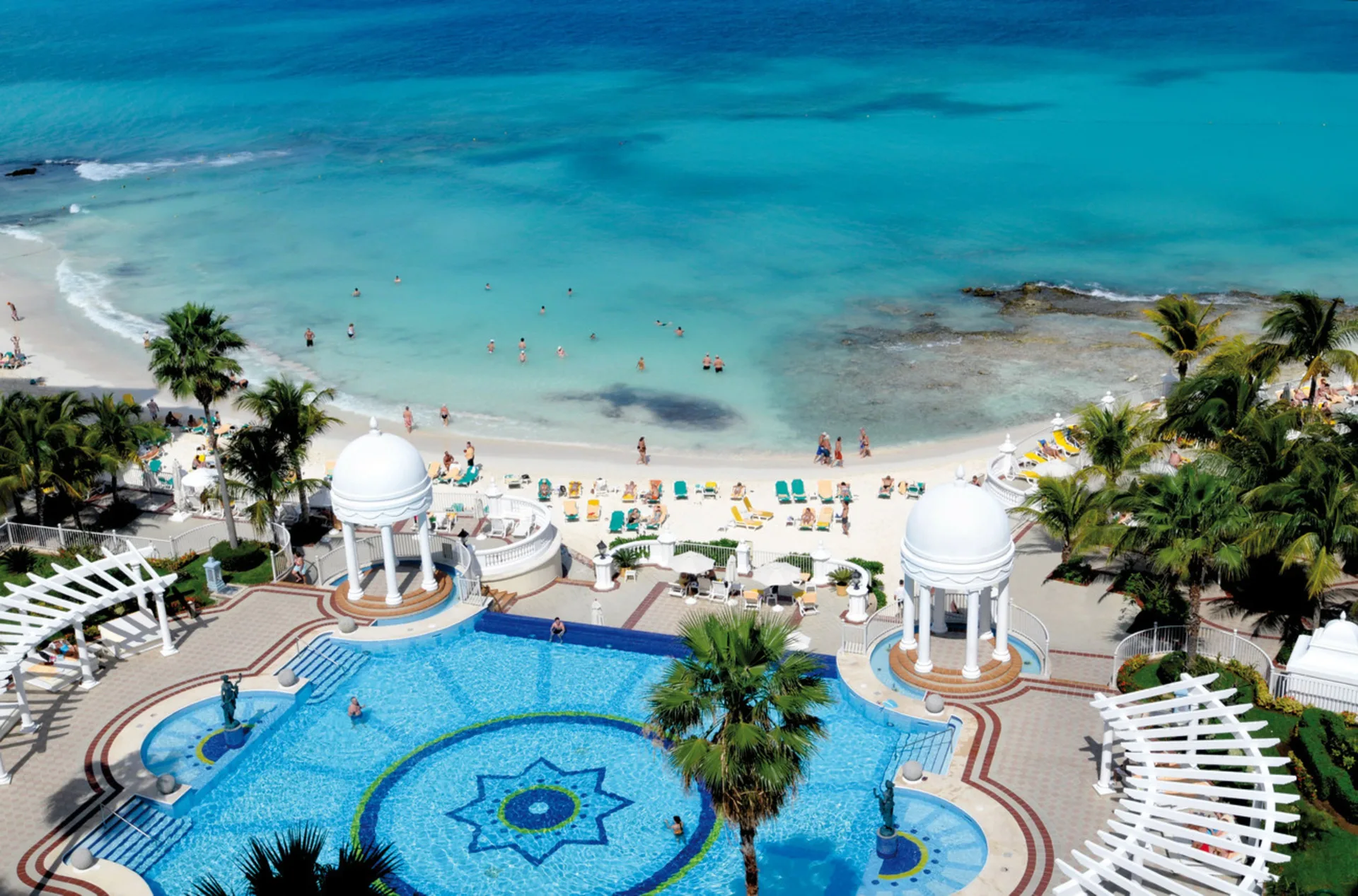Pool terraces, domed pavilions, beach loungers, and clear sea at Hotel Riu Palace Las Americas.