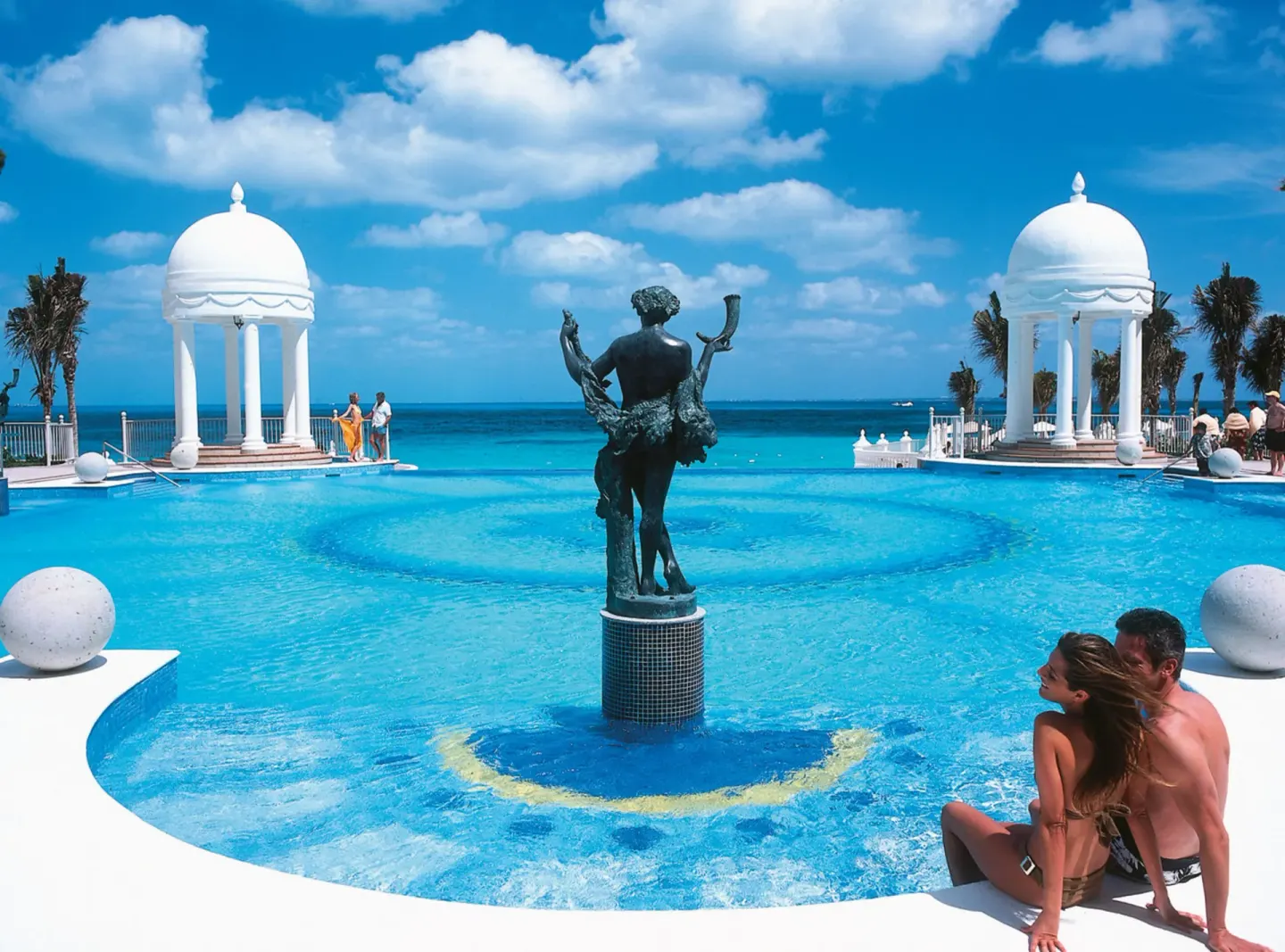 Couple by a pool fountain statue and domed pavilions at Hotel Riu Palace Las Americas.