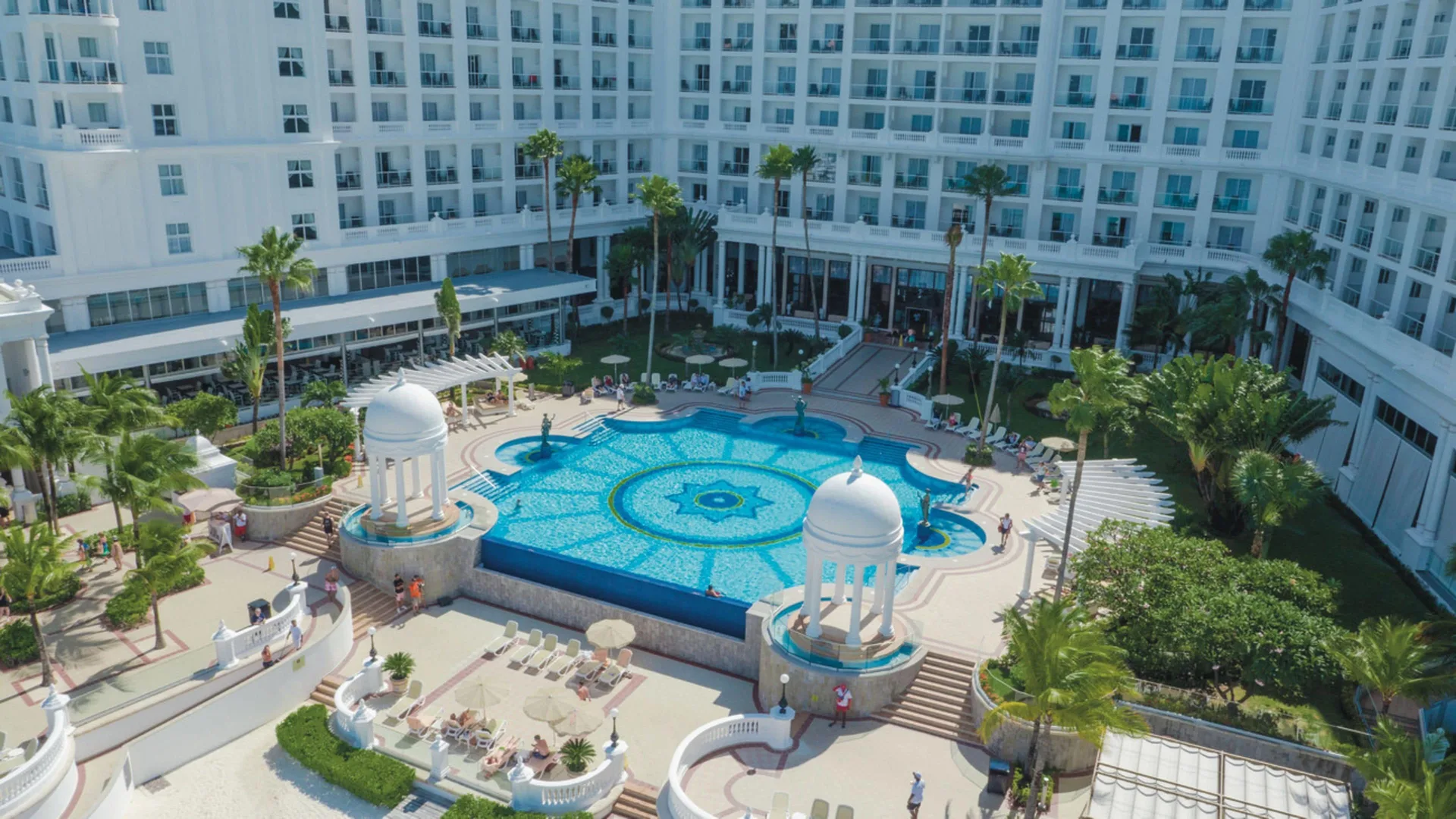 Sunny courtyard pool at Hotel Riu Palace Las Americas with loungers, palms, and white buildings.