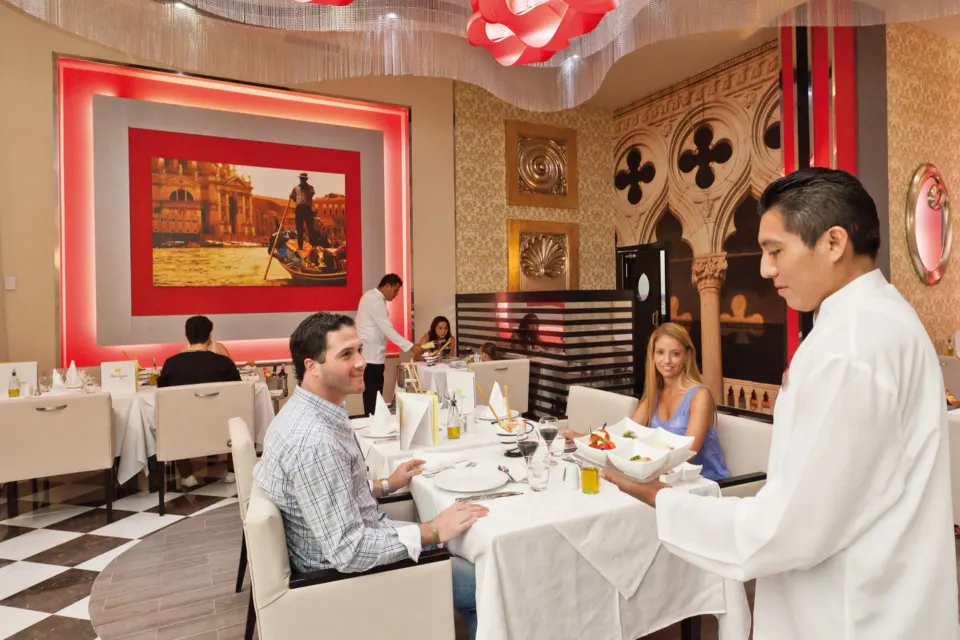 Romantic indoor Italian-style dining room with waiter serving a couple at Hotel Riu Palace Mexico