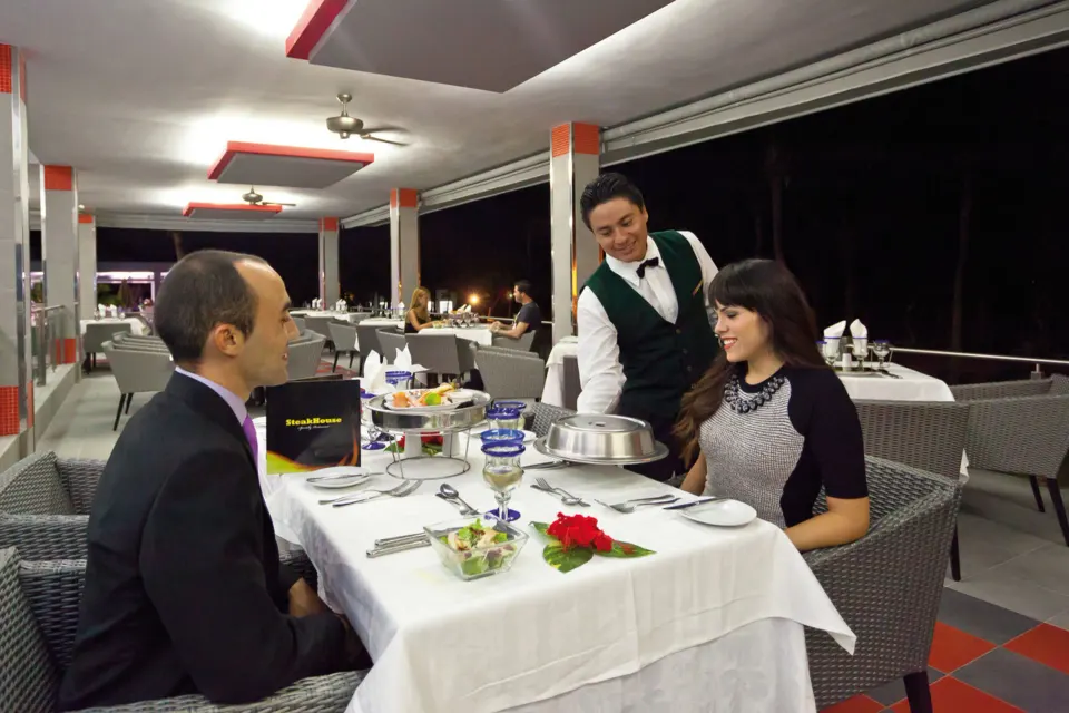 Couple dining on a covered terrace with waiter serving at night at Hotel Riu Palace Mexico