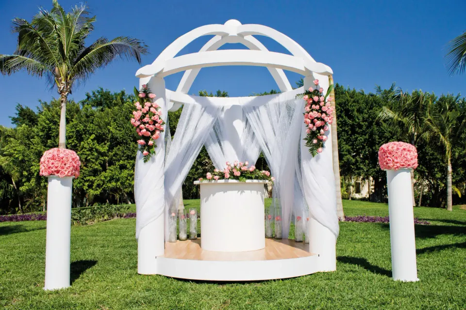 White ceremony pavilion with pink floral columns on green lawn at Hotel Riu Palace Mexico