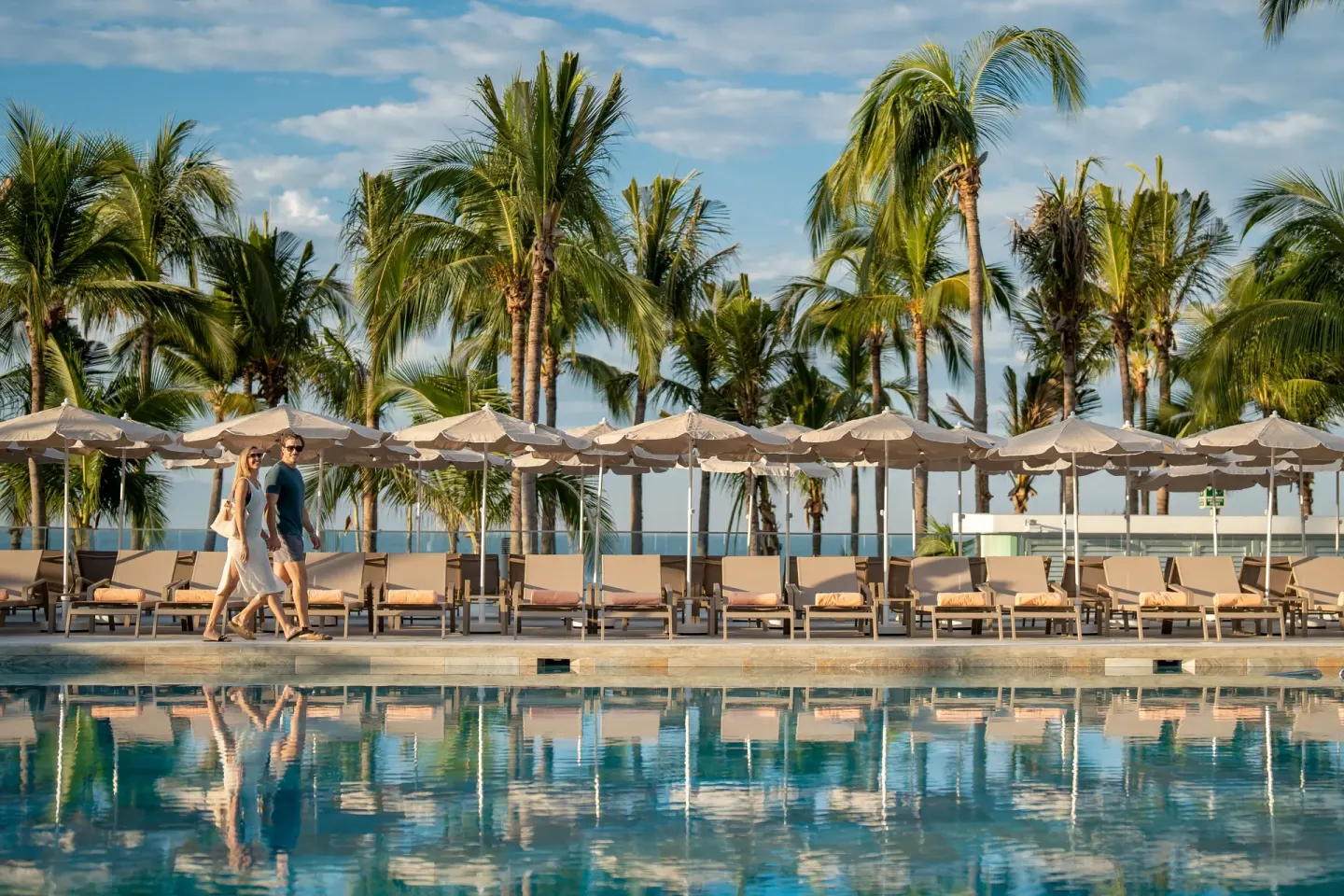 Poolside walkway with loungers, umbrellas, palm trees, and two guests at Hotel Riu Palace Pacifico.