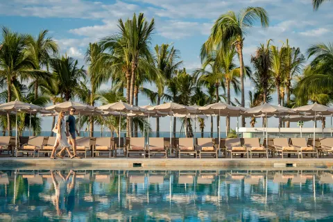 Poolside walkway with loungers, umbrellas, palm trees, and two guests at Hotel Riu Palace Pacifico.