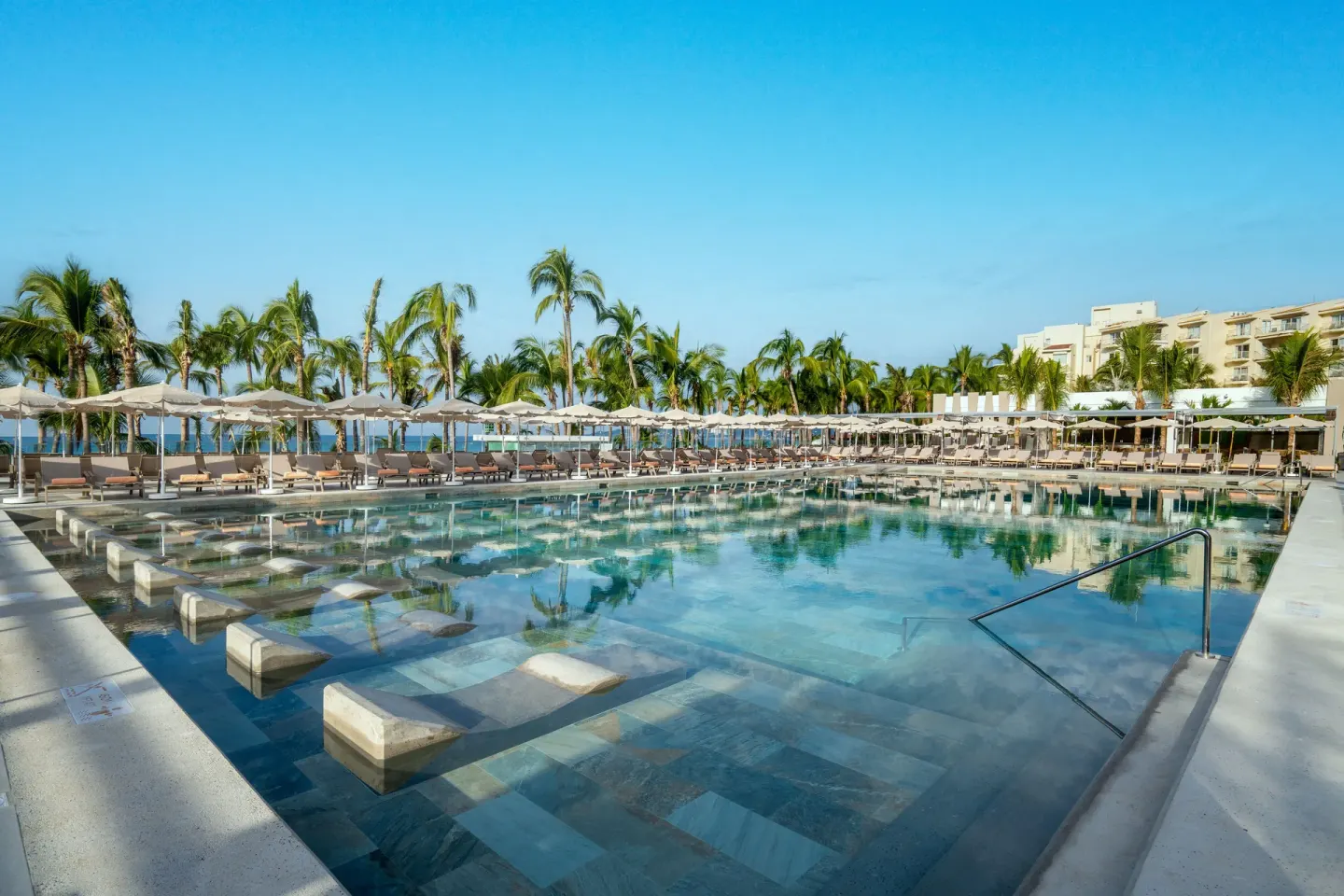 Large resort pool with loungers, umbrellas, palm trees, and ocean beyond at Hotel Riu Palace Pacifico.