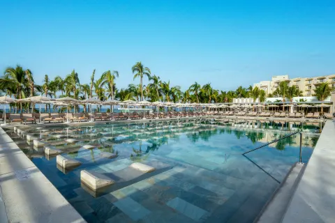 Large resort pool with loungers, umbrellas, palm trees, and ocean beyond at Hotel Riu Palace Pacifico.