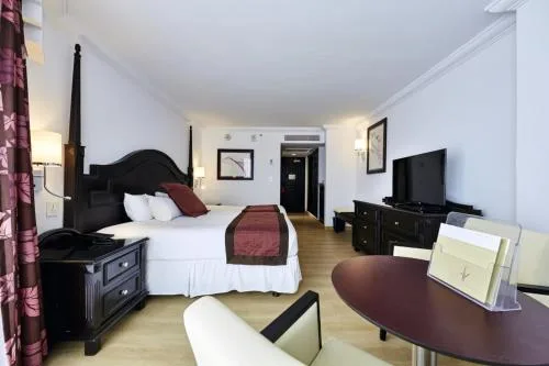 Suite bedroom with a bed, TV, round table, and entry door at Hotel Riu Palace Paradise Island.