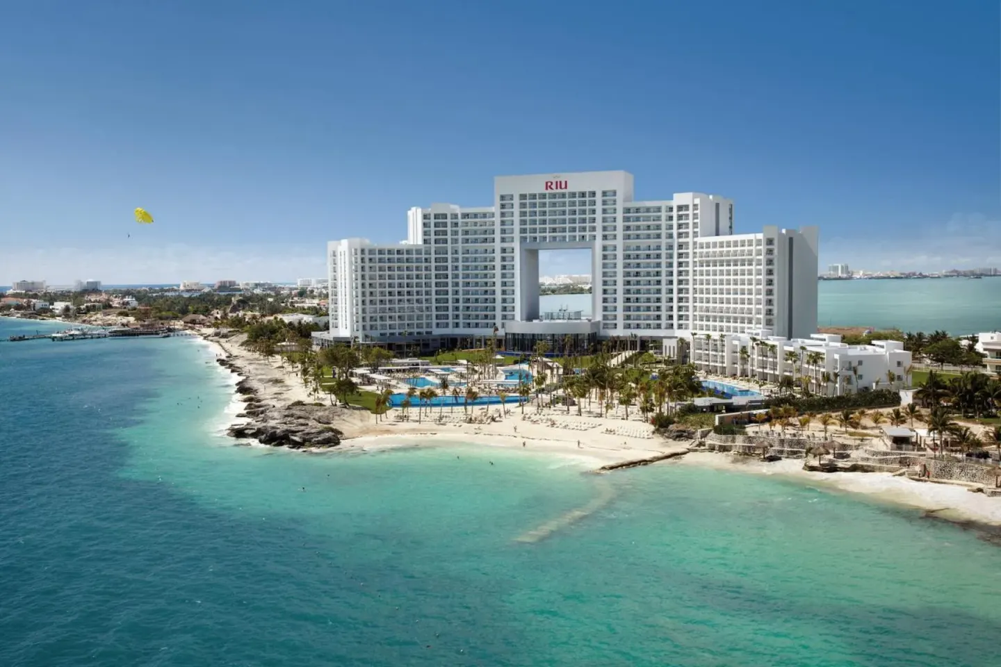 Hotel Riu Palace Peninsula rises beside turquoise water and a sandy shoreline.