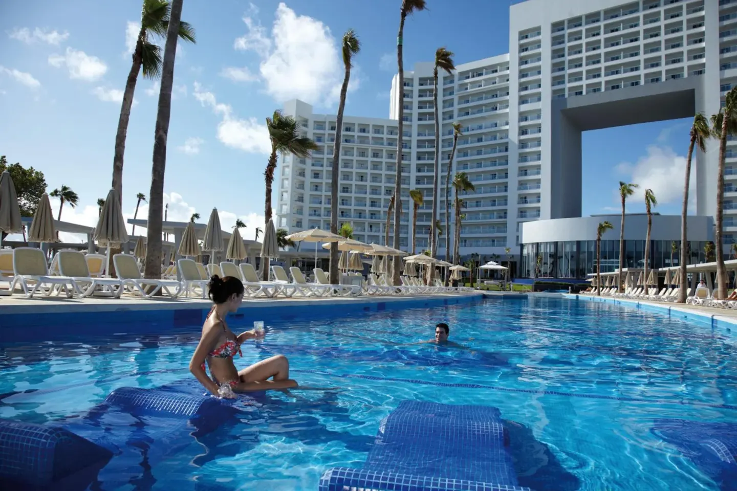 Pool with submerged loungers, palm trees, and hotel towers at Hotel Riu Palace Peninsula.