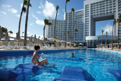 Pool with submerged loungers, palm trees, and hotel towers at Hotel Riu Palace Peninsula.