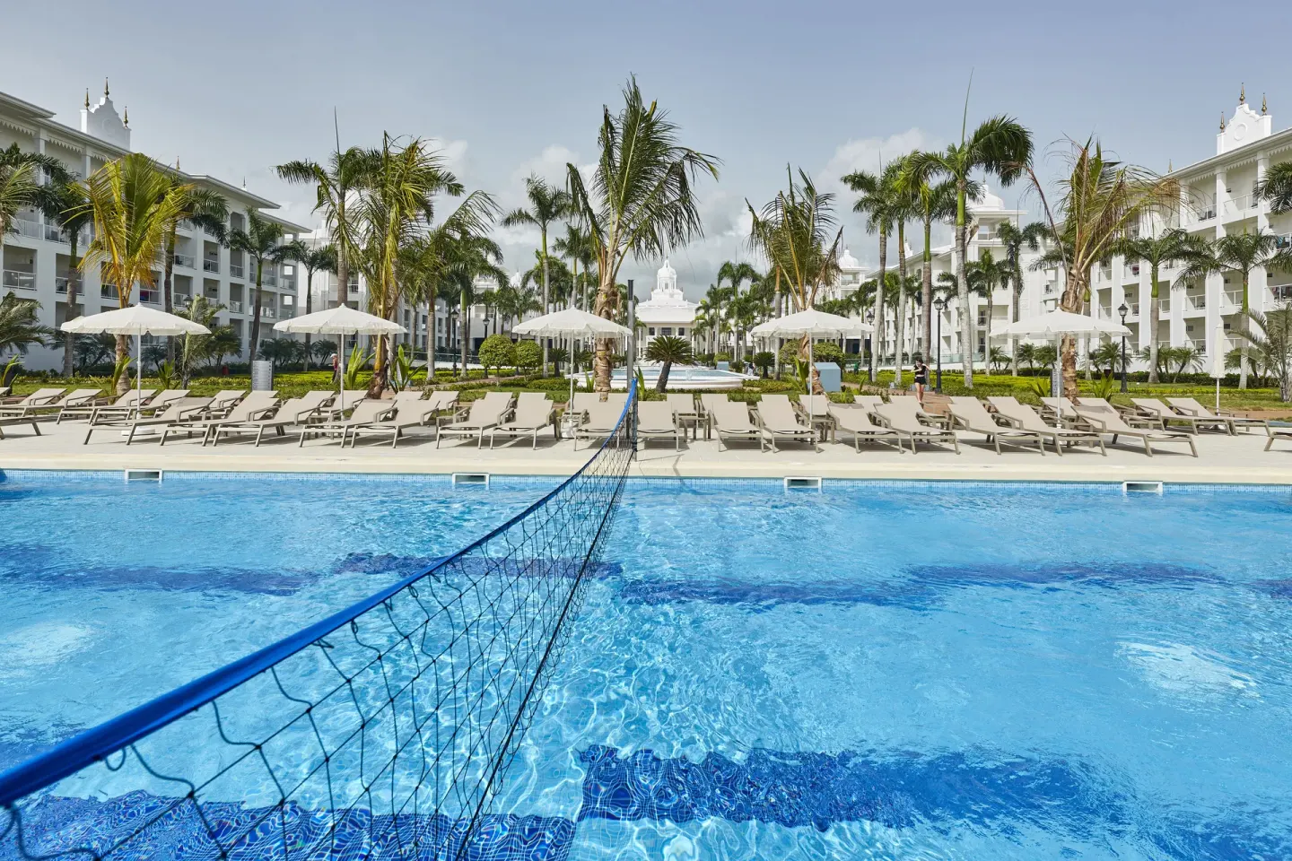 Pool volleyball net, loungers, palms, and white buildings at Hotel Riu Palace Punta Cana.