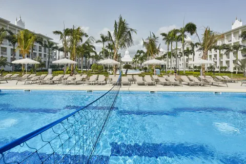 Pool volleyball net, loungers, palms, and white buildings at Hotel Riu Palace Punta Cana.