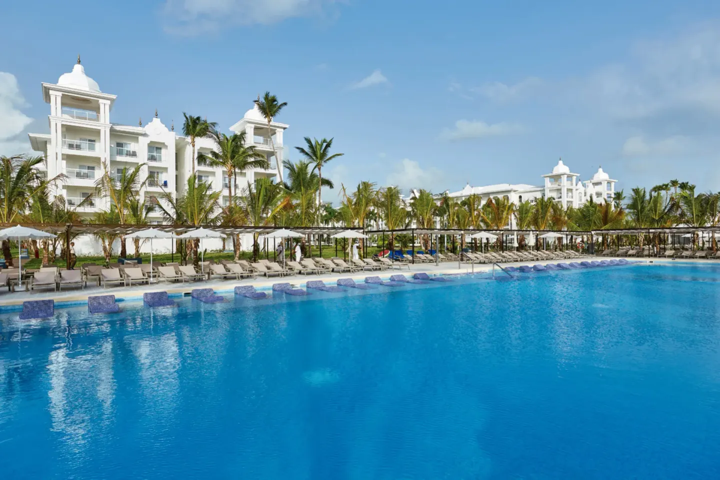 Wide resort pool with loungers, palms, and white buildings at Hotel Riu Palace Punta Cana.
