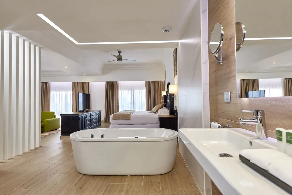 Suite bath area with freestanding tub, vanity, king bed, and lounge beyond at Hotel Riu Palace Punta Cana.