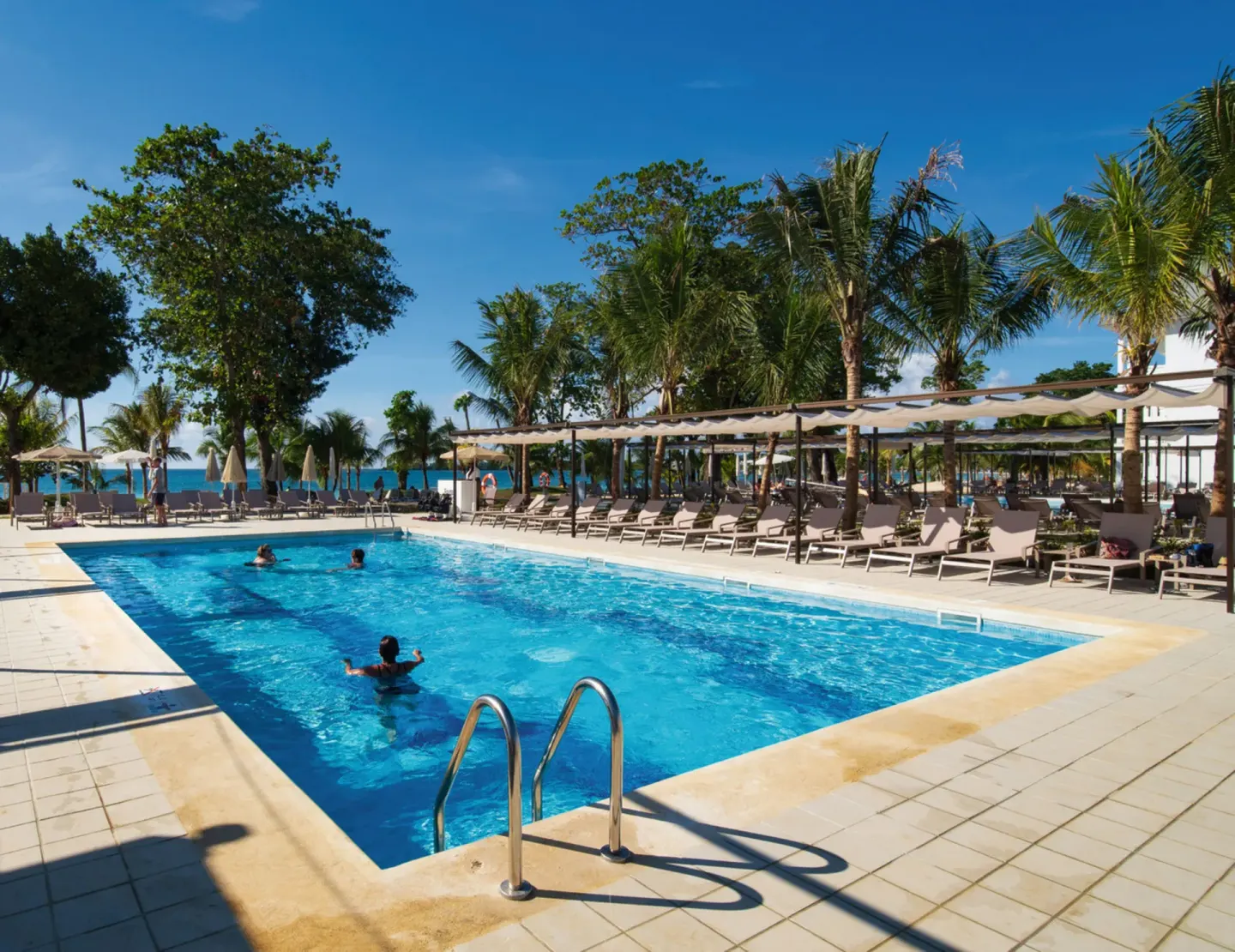Pool with metal ladder, palms, and shaded loungers at Hotel Riu Palace Tropical Bay.