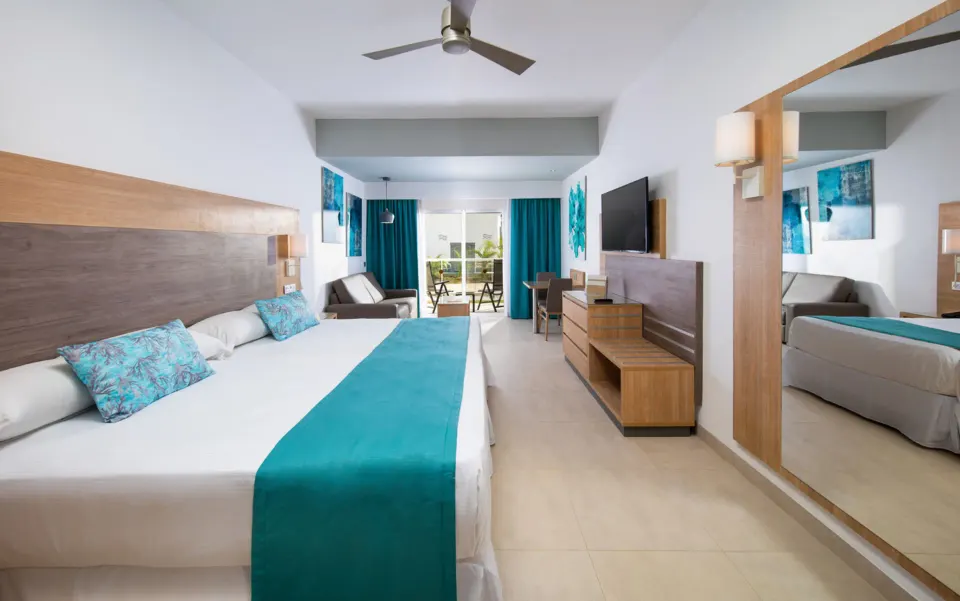 Junior Suite Garden View with bed, mirror, TV console, seating area, and balcony doors at Hotel Riu Palace Tropical Bay.