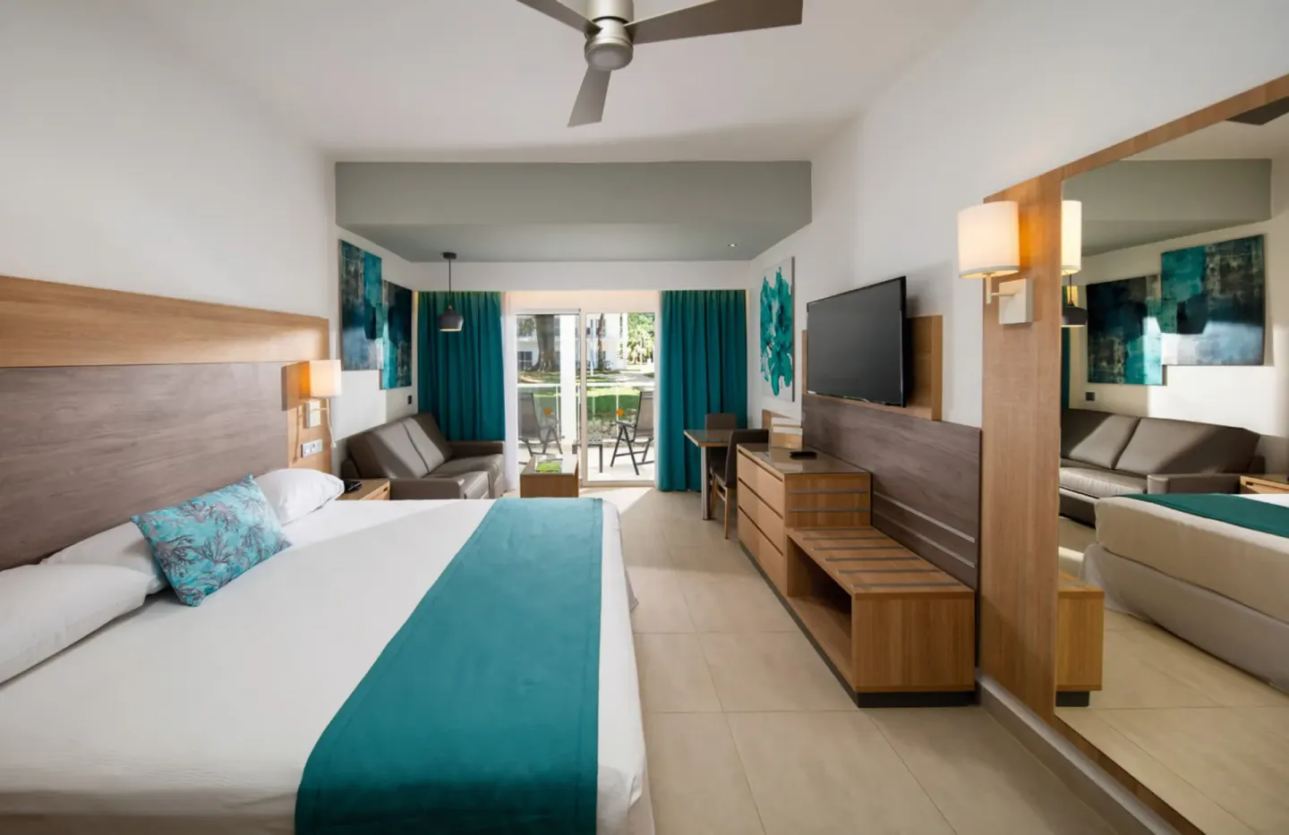 Standard Room with bed, sofa, TV, mirror and balcony at Hotel Riu Palace Tropical Bay.