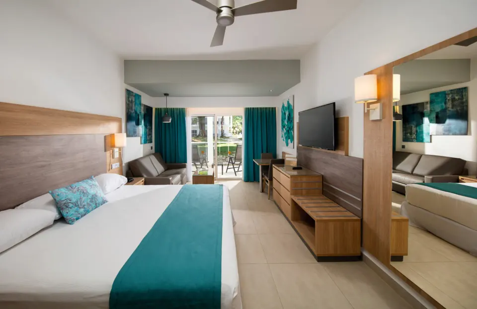 Standard Room with bed, sofa, TV, mirror and balcony at Hotel Riu Palace Tropical Bay.
