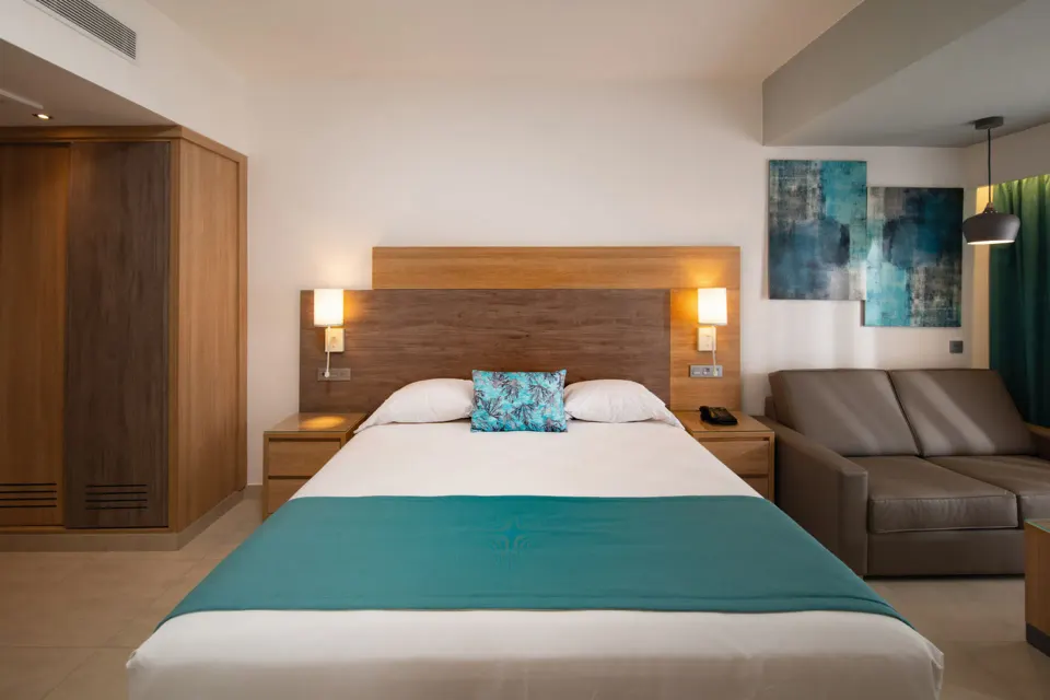 Standard Room bed with sofa, wardrobes and wall art at Hotel Riu Palace Tropical Bay.