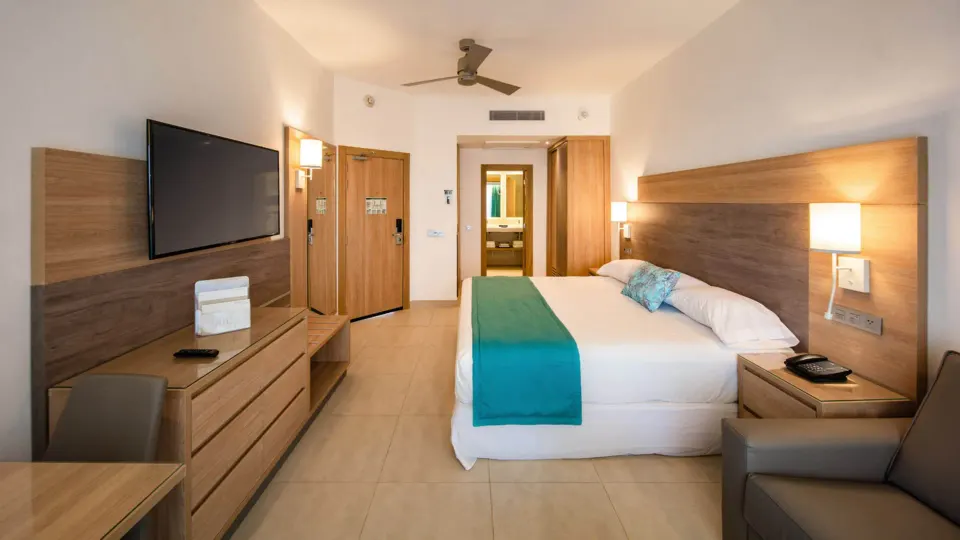 Standard Room with bed, TV, desk, sofa and bathroom doorway at Hotel Riu Palace Tropical Bay.