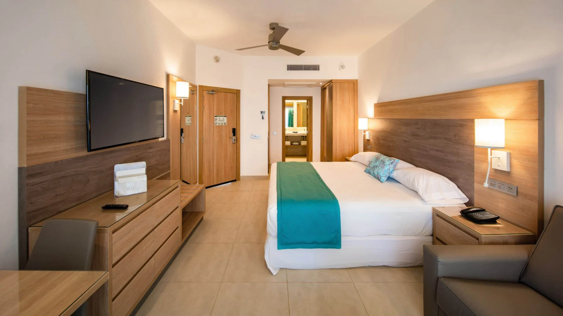 Standard Room with bed, TV, desk, sofa and bathroom doorway at Hotel Riu Palace Tropical Bay.
