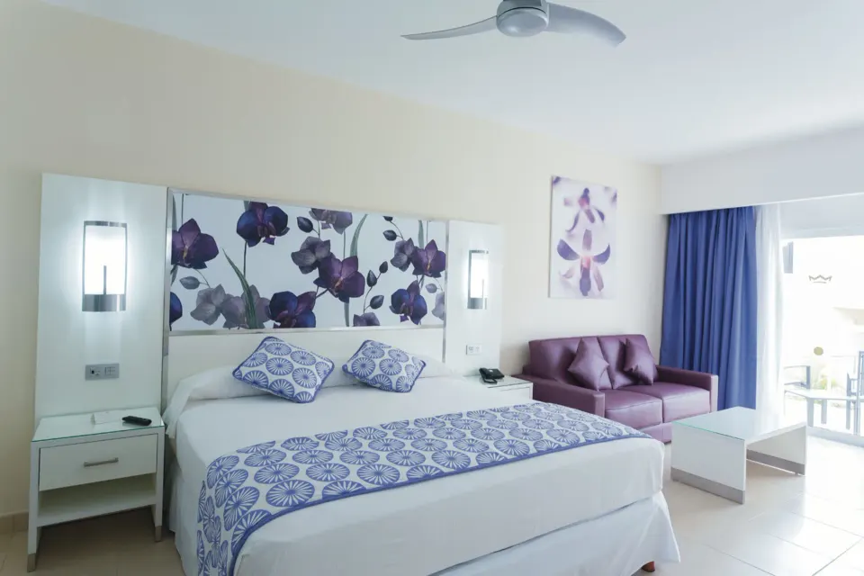 King bed with purple sofa and wall art in a bright suite at Hotel Riu Playacar