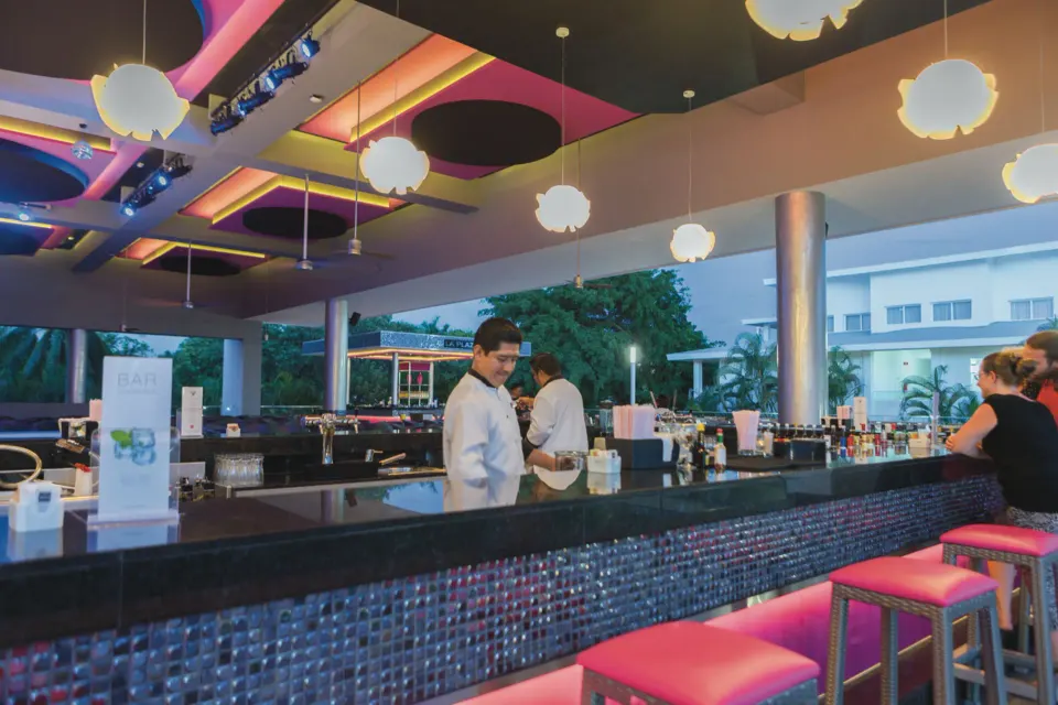 Long bar counter with pink stools and staff serving drinks at Hotel Riu Playacar