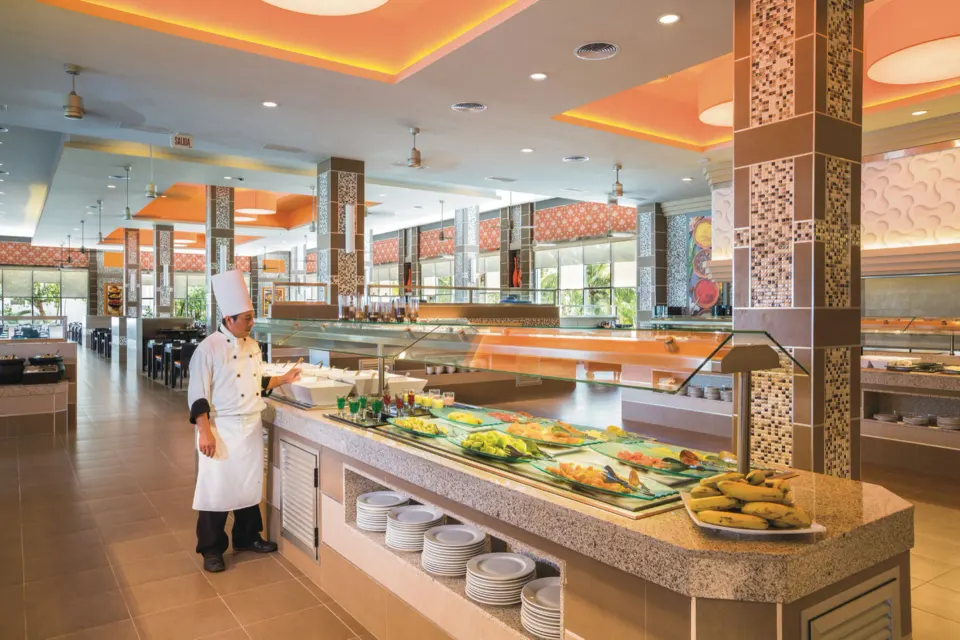Spacious buffet hall with islands and a chef standing near service counters at Hotel Riu Playacar