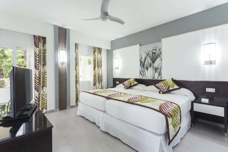Angled view of twin beds, TV and balcony windows in a Junior Suite at Hotel Riu Playacar