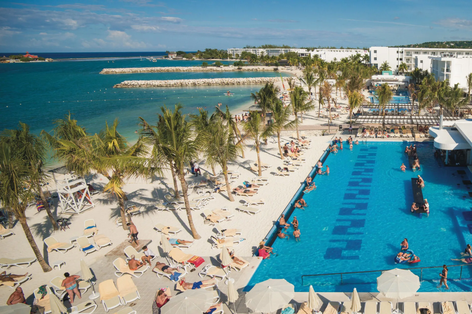 Aerial view of Hotel Riu Reggae pool and beach with loungers, palms, and calm bay water.