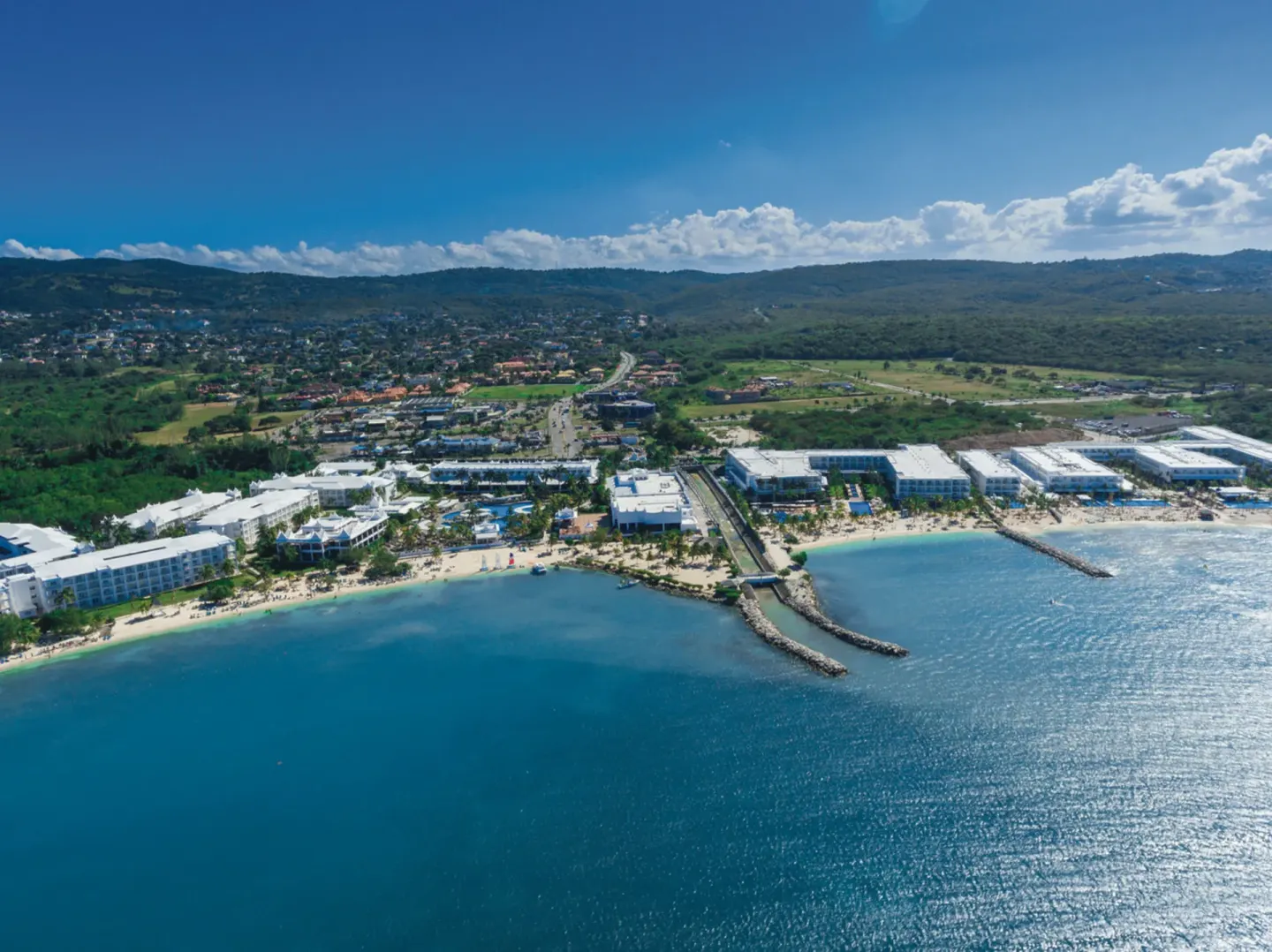 Distant aerial view of Hotel Riu Reggae along the bay with white buildings and rocky breakwaters.
