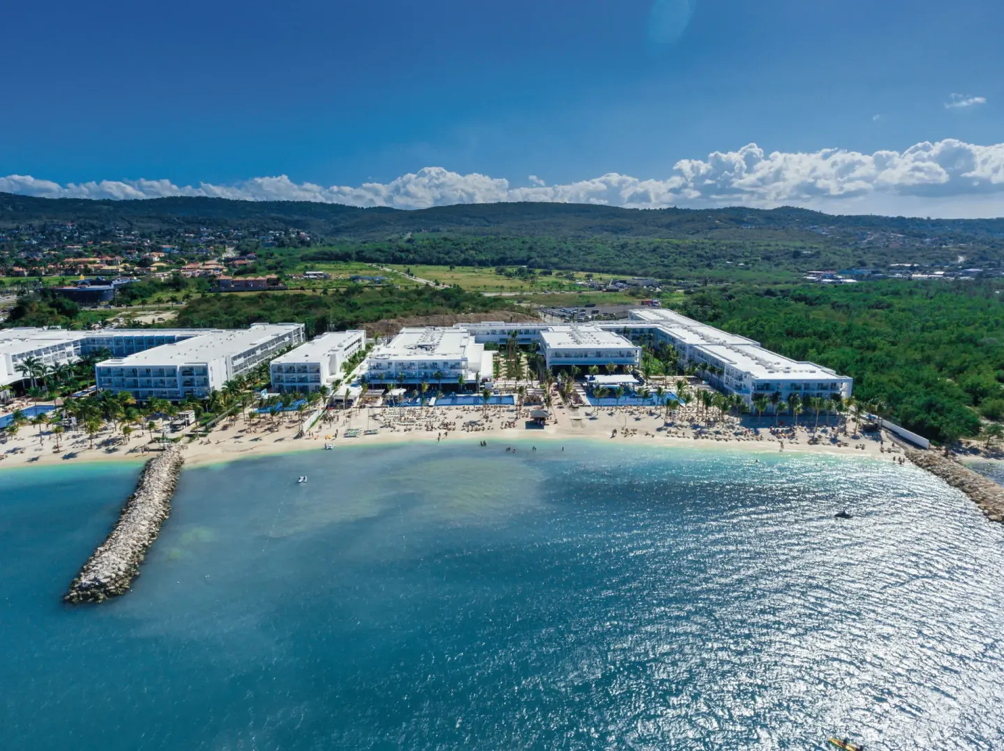 Aerial view of Hotel Riu Reggae beachfront with pools, loungers, a breakwater, and open sea.