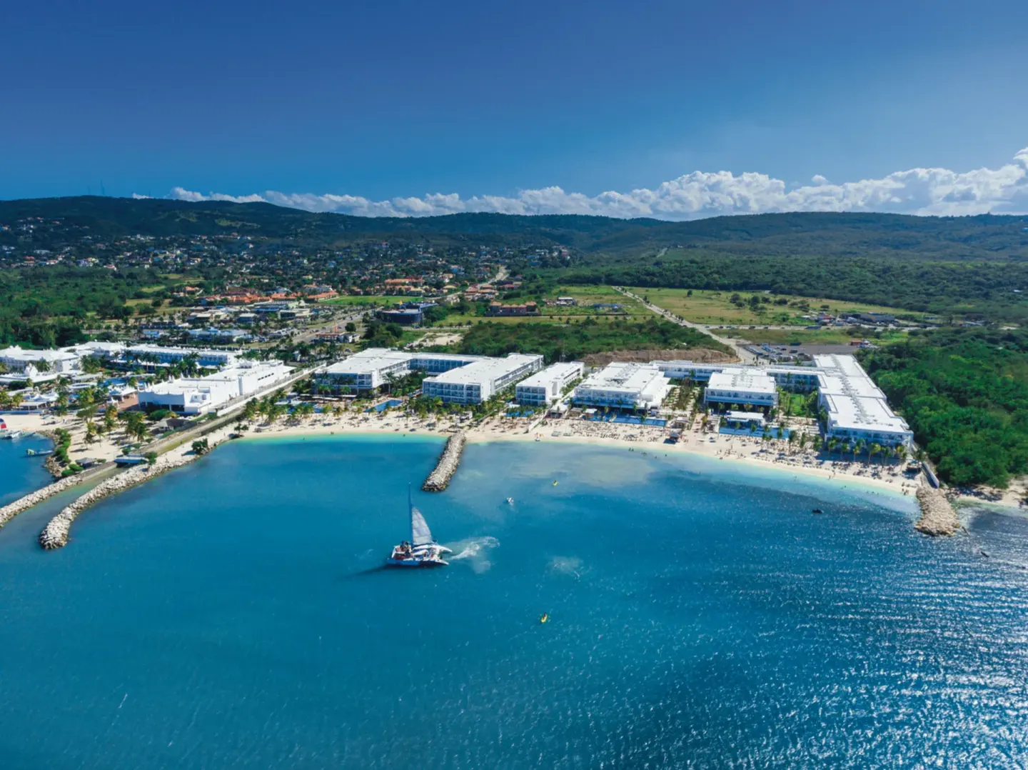 Aerial coastline view of Hotel Riu Reggae with breakwaters, beach, and a catamaran offshore.