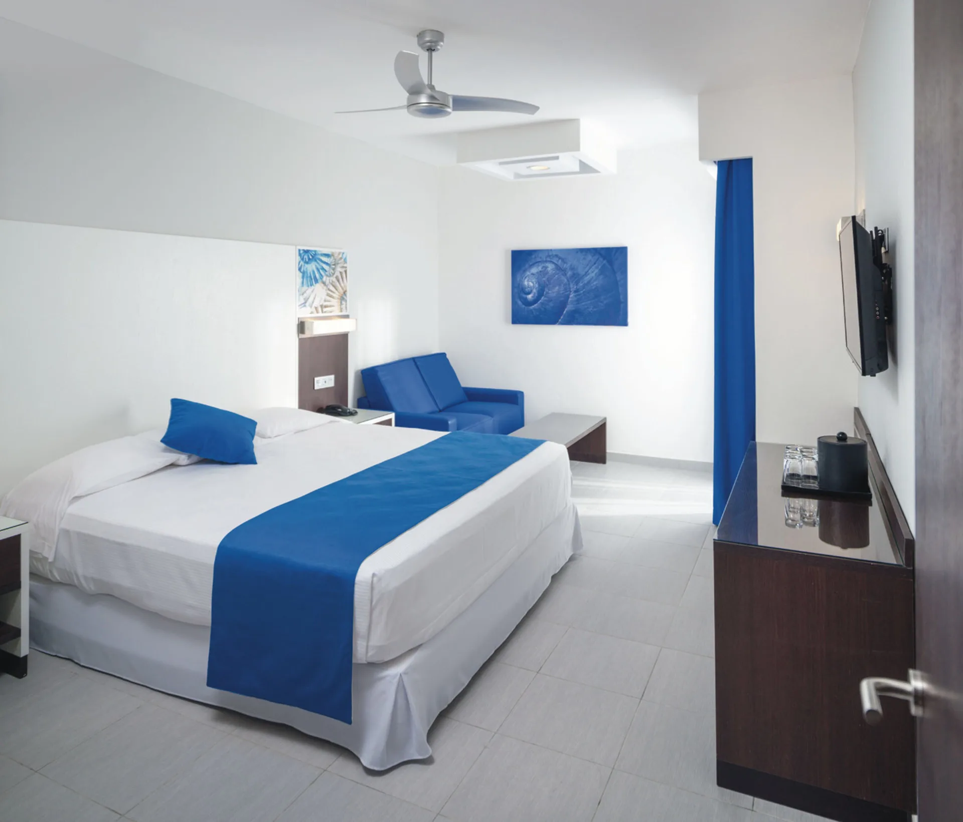 Guest room at Hotel Riu Reggae with king bed, blue sofa, wall-mounted TV, and desk.
