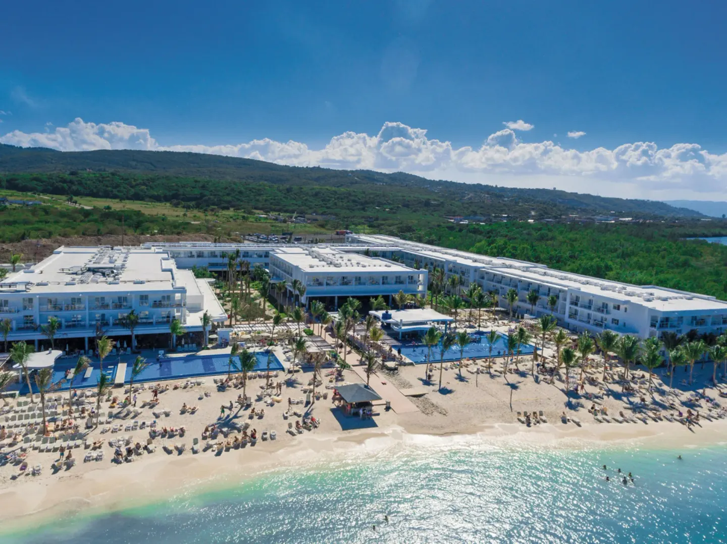 Hotel Riu Reggae seen from above with pools, beach loungers, palm trees, and hills behind.