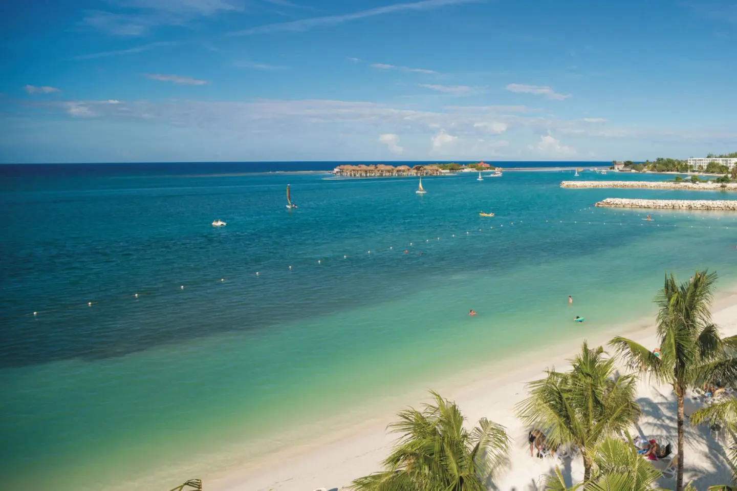 Beach view near Hotel Riu Reggae with turquoise water, palms, swimmers, and small sailboats.
