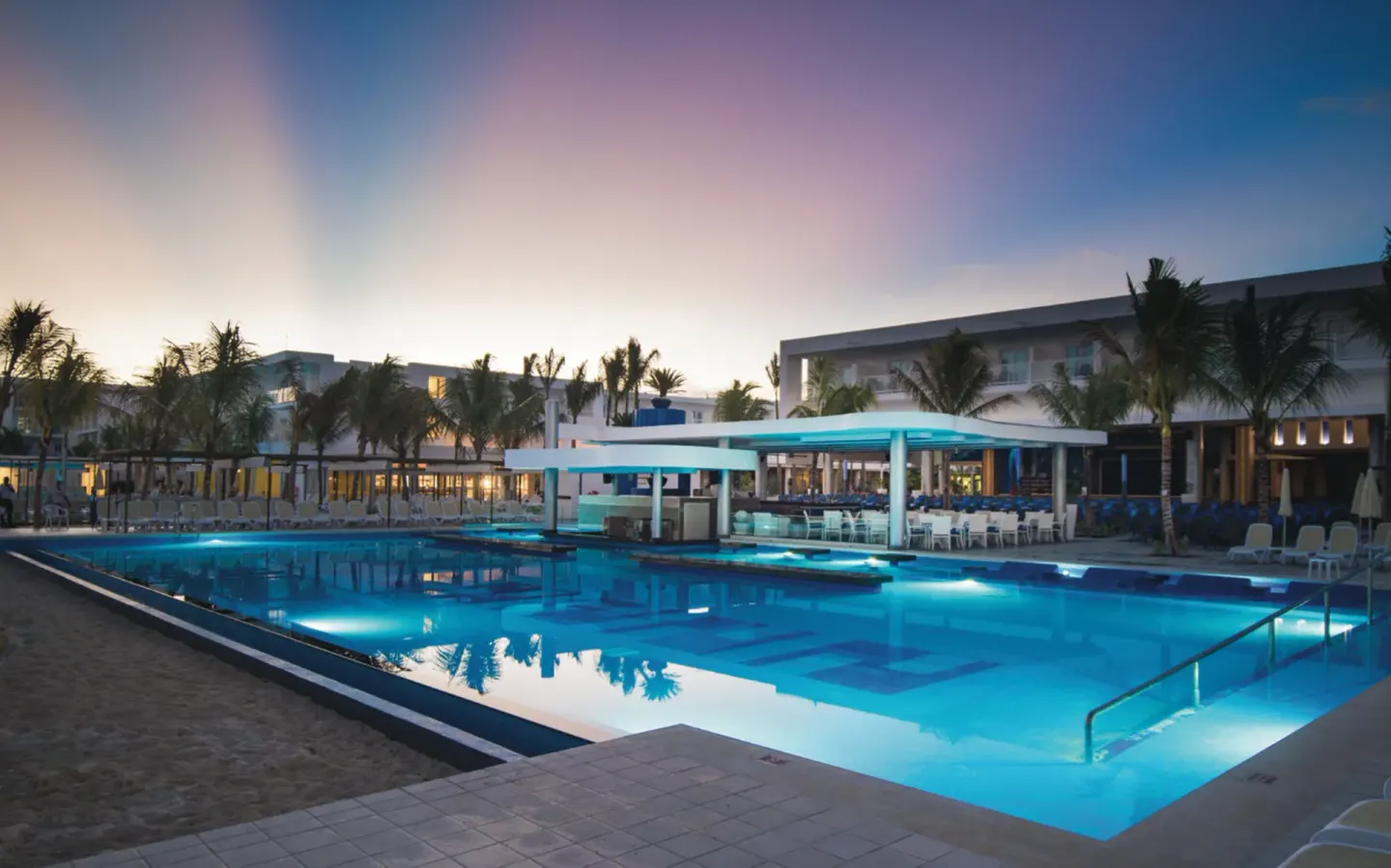 Twilight pool scene with a bar pavilion and lit water at Hotel Riu Reggae.