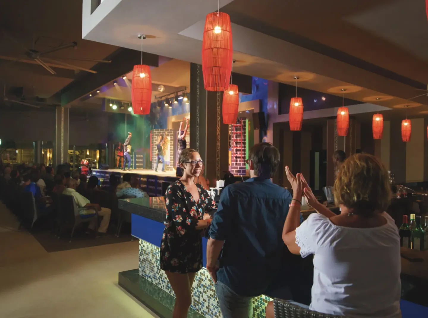 Guests stand by a bar while a stage show plays in the background at Hotel Riu Reggae.