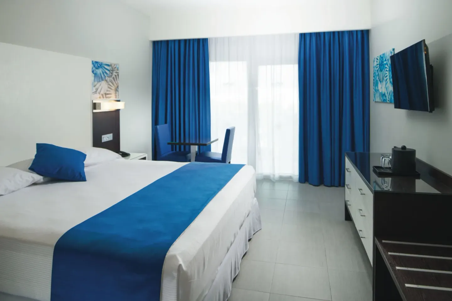 Tropical view room with one bed, blue curtains, TV and seating area at Hotel Riu Reggae.
