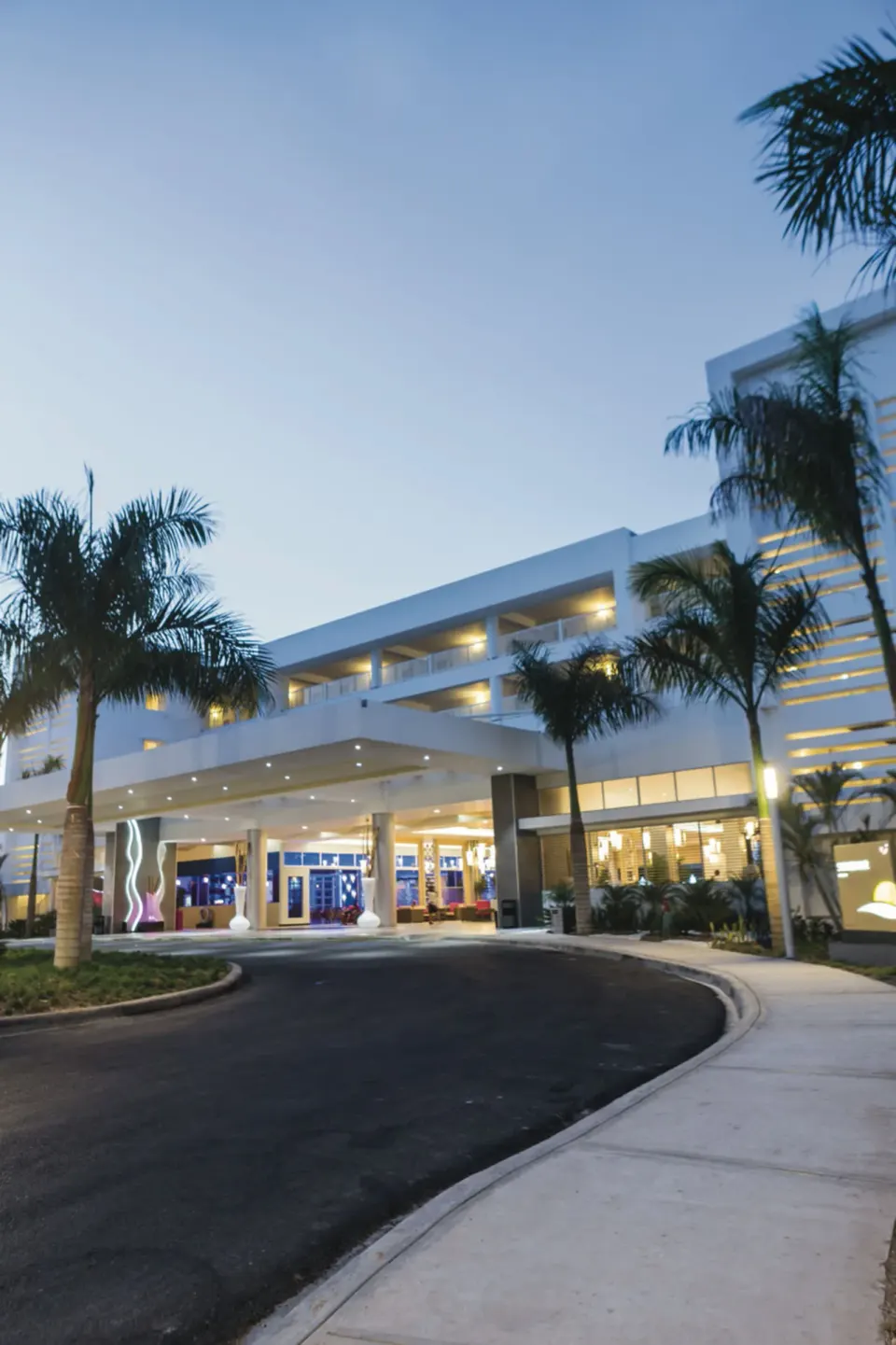 Curved driveway and illuminated entrance canopy at Hotel Riu Republica framed by palm trees at dusk.