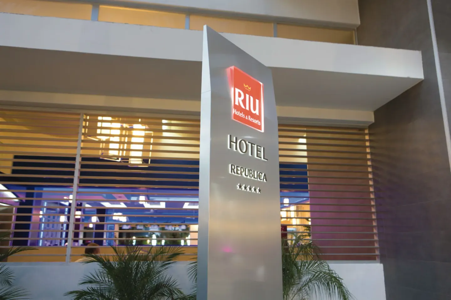 Illuminated entrance sign outside Hotel Riu Republica with palms and lobby windows behind it.