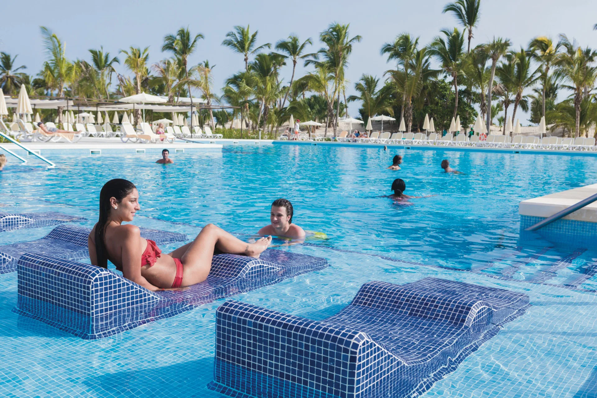 In-water lounge chairs and swimmers in a palm-lined pool at Hotel Riu Republica.