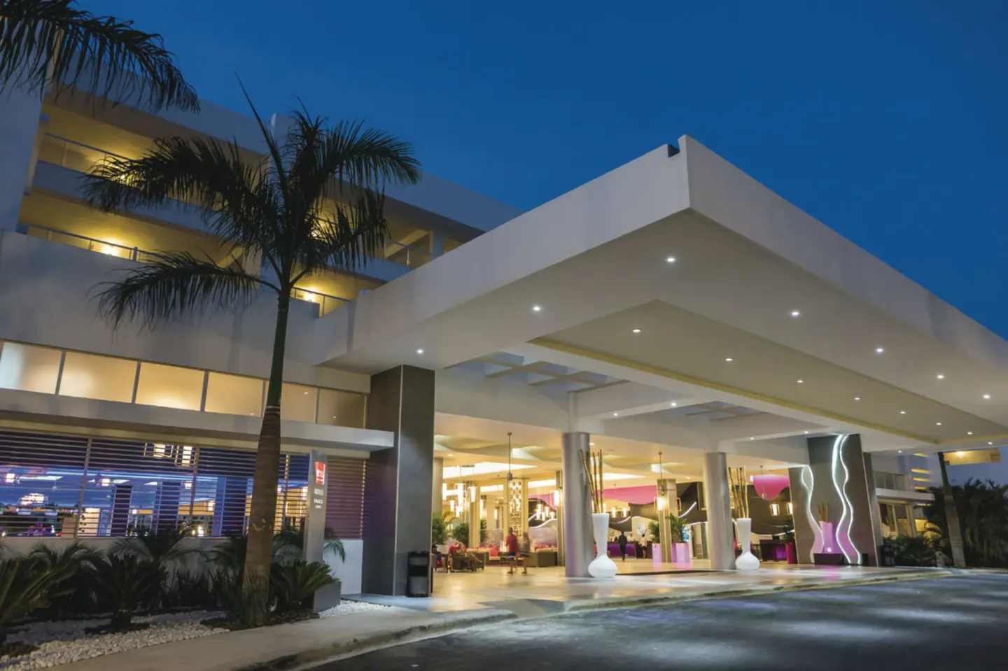 Covered nighttime entrance at Hotel Riu Republica with palm trees and a glowing lobby beyond.