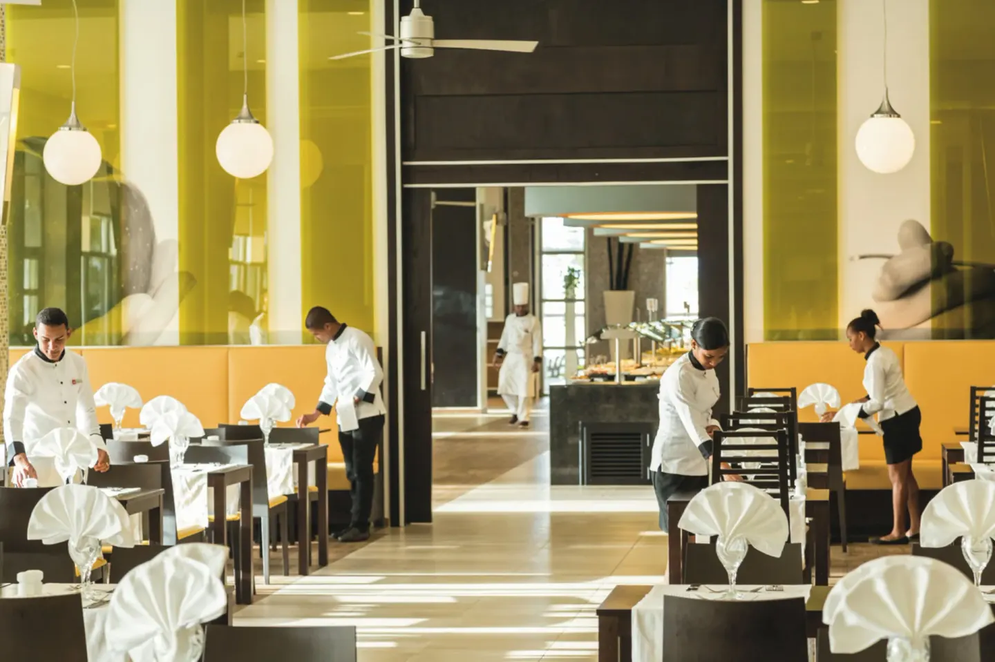 Restaurant staff set tables in a yellow-accented dining room at Hotel Riu Republica.